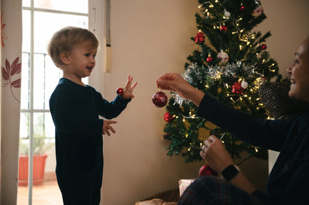 A toddler shares a festive moment with a parent as they hang red ornaments on a lit Christmas tree, capturing warmth, joy, and togetherness in a cozy holiday living room.の写真素材