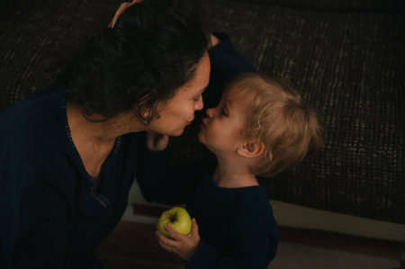 A mother leans in to kiss her toddler who holds an apple. The moment captures family bonding, care, and everyday affection in a cozy home setting.の写真素材