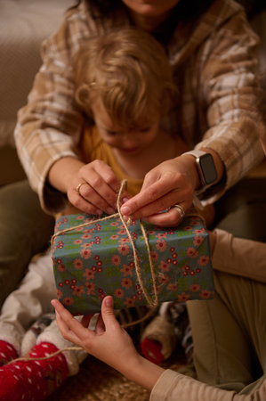 Warm family moment as hands tie a jute string on a floral gift while a child watches, cozy, festive...