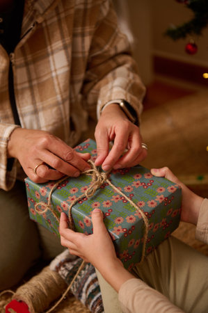 Hands tie a rustic twine bow on a blue floral gift box, assisted by a plaid-clad person. Warm, festive moment of gifting and holiday cheer.の写真素材