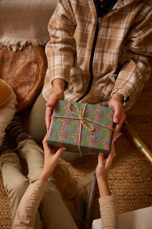 Two friends share a wrapped blue floral gift on a woven rug, wearing plaid jackets. The scene feels warm, festive, and intimate, capturing a simple holiday moment of giving and joy.の写真素材