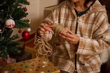A person in a plaid jacket ties jute twine to a gift beside a decorated Christmas tree. Soft lights and colorful wrapping create a warm, homey holiday scene.の写真素材