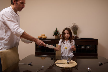 A father hands a plate to his young daughter during a meal at the dining room. They share a calm, domestic moment as they prepare and enjoy a simple family supper.の写真素材