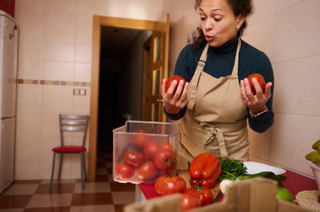 A woman in a beige apron stands in a bright kitchen, holding ripe tomatoes while a clear box of...
