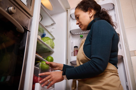 A home cook in a teal sweater and beige apron selects fresh fruit from an open fridge, highlighting everyday kitchen duties and healthy eating.の写真素材