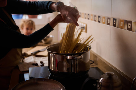 Close-up of hands stirring spaghetti into a pot on the stove, with a child in the background. Warm, cozy home cooking scene capturing family moments.の写真素材