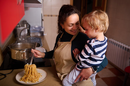 Warm, smiling mother cooks spaghetti on the stove while holding her young son, capturing family bonding, cooking together, and cozy home life in a kitchen.の写真素材