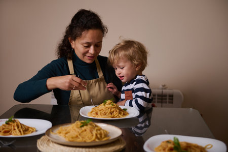 A smiling mother in an apron shares spaghetti with her young child at a glass table, capturing a warm, intimate family moment of togetherness and home cooking.の写真素材