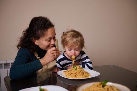 A loving mother feeds her toddler as they share a plate of spaghetti, capturing a tender family moment during a home meal.の写真素材
