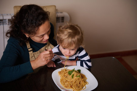A mother feeds her child spaghetti at a cozy kitchen table, capturing a warm moment of care, family, and daily life.の写真素材