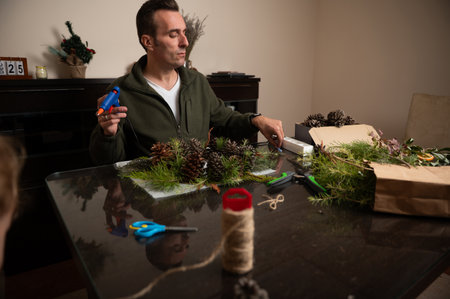 A man decorates festive Christmas decorations using pine cones, pine branches, twine, and glue on a dining table in a cozy home setting, conveying warmth, creativity, and holiday craft.の写真素材