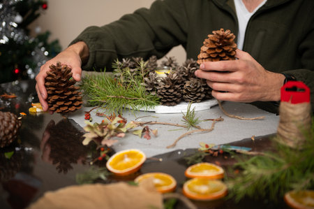 A person crafts pine cone decorations with twine and greenery at a cozy home table, capturing festive, handmade holiday spirit and togetherness.の写真素材