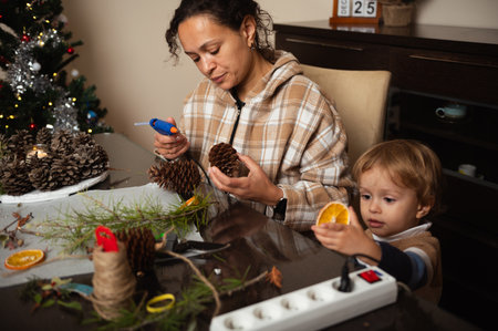 A woman and child craft festive decorations with pinecones, twine, and oranges at a cozy table. Warm light, Christmas tree, and crafting create a welcoming family moment.の写真素材
