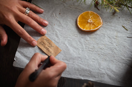 A close view shows hands writing a brown tag with Merry Christmas , while a fresh orange slice and...