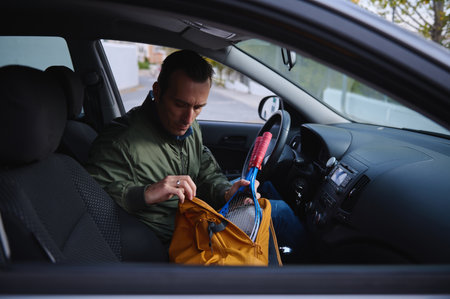 A man sits in a car, opening a yellow backpack to access gear, suggesting travel, prep, and outdoor activity.の写真素材
