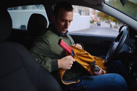 A man sits in a car, packing a bright orange backpack with a pink umbrella and water bottle. The scene conveys travel, preparation, and casual outdoor activity.の写真素材