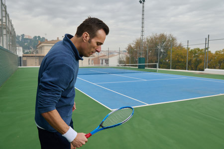 A man on a blue tennis court holds a racket, ready to play. He wears a blue fleece and wristband under an overcast sky, with a fenced backdrop.の写真素材