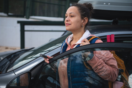 A woman stands beside an open car door, holding a phone and wearing a backpack with hiking gear. She...