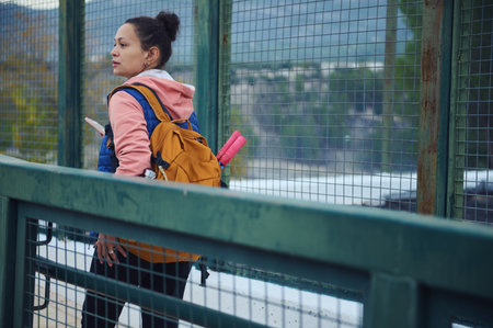 A woman wearing a pink hoodie and orange backpack stands on a bridge, looking into the distance with...