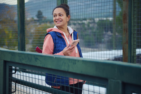 A cheerful woman stands on a railing bridge outdoors, holding a smartphone, wearing a pink hoodie and blue vest, enjoying a moment of casual tech use.の写真素材