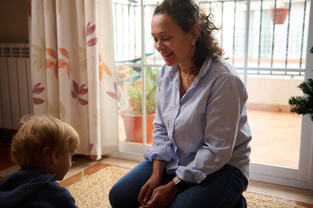 A smiling mother sits on the floor with a young child inside a bright home, sharing a warm moment by...