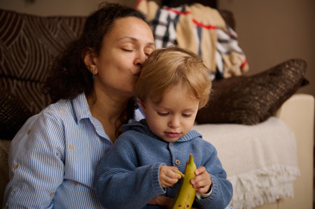 A loving mother sits with her toddler at home, sharing a kiss while the child examines a banana. Warm, candid family moment highlighting care, bonding, and simple joys.の写真素材