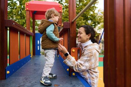 A mother kneels beside her toddler on a colorful playground, sharing a loving, protective moment...