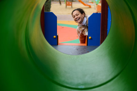 A smiling mother peers through a green playground tunnel toward her child, capturing joyful outdoor...