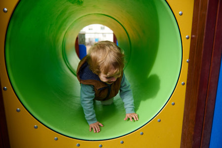 A young child crawls through a bright green playground tunnel, smiling as they explore. The scene captures playful moments of childhood, curiosity, and safe, supportive play in a park setting.の写真素材