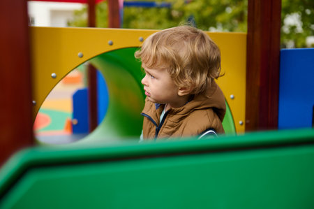 A toddler in a brown jacket stands at a colorful playground, watching intently as autumn colors surround him. A mother's care and playful environment convey warmth, safety, and carefree childhood.の写真素材
