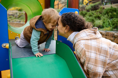 A young child crawls toward a smiling mother on a colorful playground. They share a warm, loving moment in a park, capturing care, happiness, and the joys of childhood.の写真素材
