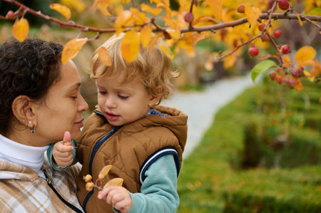 A mother and her young child enjoy a peaceful autumn day in a park, touching leaves and sharing a...