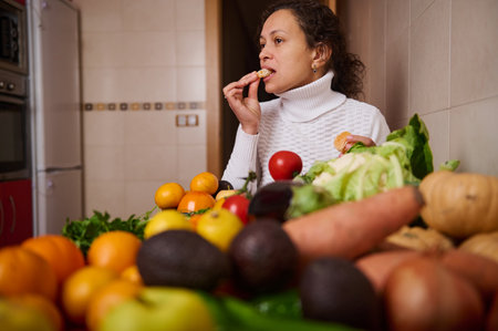 A woman in a white turtleneck snacks amid a colorful spread of fruits and vegetables in a kitchen,...