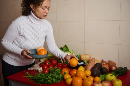 A woman in a white turtleneck selects oranges among a vibrant spread of fresh produce - tomatoes, greens, and root vegetables - on a red counter, highlighting home cooking.の写真素材