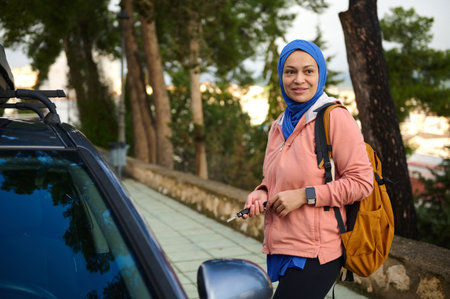A confident woman wearing a blue hijab and peach jacket stands beside a car, carrying a backpack and keys, ready for travel through a city street.の写真素材