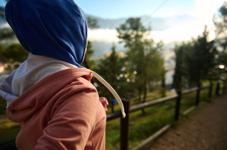 A Muslim woman wearing a blue hijab and pink hoodie walks along a park path, with blurred trees and a fence in the background, conveying everyday life and calm.の写真素材