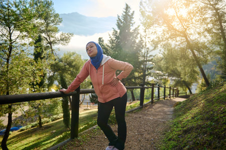 A Muslim Arab woman wearing a blue hijab and peach jacket leans on a wooden railing along a sunlit forest path in Europe, capturing a calm, healthy outdoor moment.の写真素材
