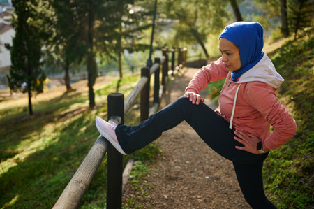 A woman in a blue hijab and pink hoodie stretches beside a wooden fence in a sunlit park, conveying fitness, balance, and everyday active life.の写真素材