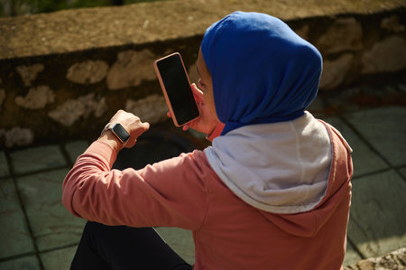 A woman in a blue hijab sits outdoors in a hoodie, focused on her smartphone. A smartwatch rests on her wrist in a casual, modern European setting.の写真素材