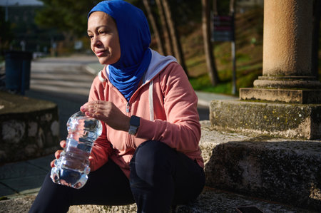 A Muslim Arab woman in a blue hijab sits on stone steps outdoors, holding a water bottle during a...