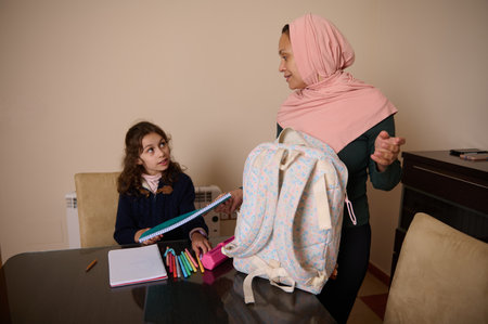 A Muslim mother in a pink hijab speaks with her daughter at a dining table, organizing school supplies beside a pastel backpack and notebooks.の写真素材