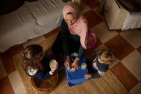 A mother in a pink hijab shares a simple meal with her two children, creating warmth, family bonds, and everyday life moments in a cozy home setting.の写真素材