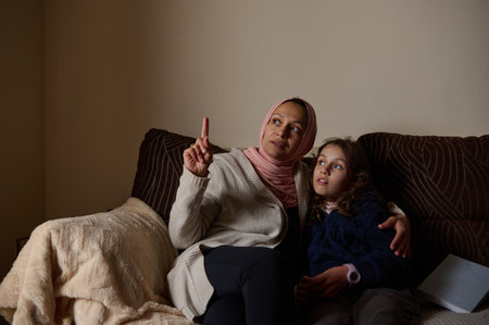 A woman in a hijab sits with her daughter on a cozy sofa, pointing upward as they share a warm, intimate moment in a bright living room.の写真素材