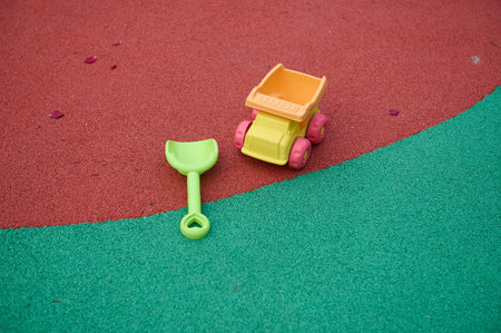 Bright green shovel and small yellow toy dump truck lie on a red and green playground surface,...