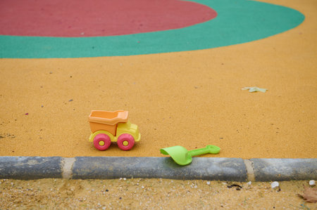 Bright orange toy dump truck and green shovel rest on a sandy playground, with a curved colorful track in the background, evoking playful outdoor fun.の写真素材