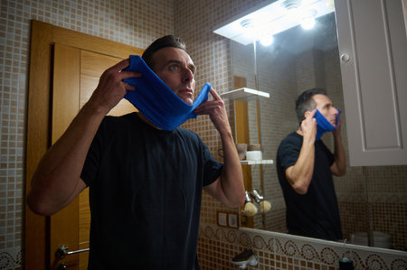 A man in a black shirt adjusts a blue towel around his face, gazing at his reflection in a tiled bathroom, capturing a candid moment of grooming and daily hygiene.の写真素材