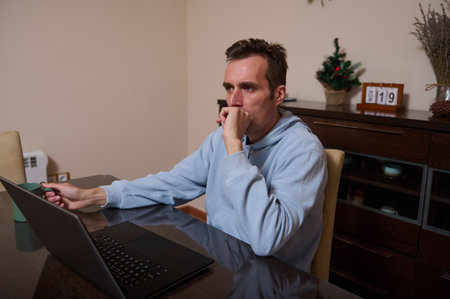A man in a light blue hoodie sits at a dining table, focused and reflective while working on a...