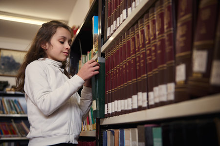A young girl in a white hoodie browses shelves of books in a school library, exploring knowledge and learning during library day.の写真素材