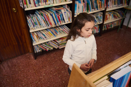 A young girl stands in a quiet library aisle, browsing books. She wears a white jacket and concentrates on selecting reading material, capturing curiosity, study habits, and the calm of learning spaces.の写真素材