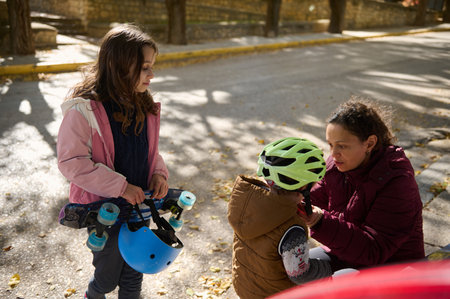 A girl in a pink jacket stands with a blue skateboard as a friend wearing a green helmet sits with a...